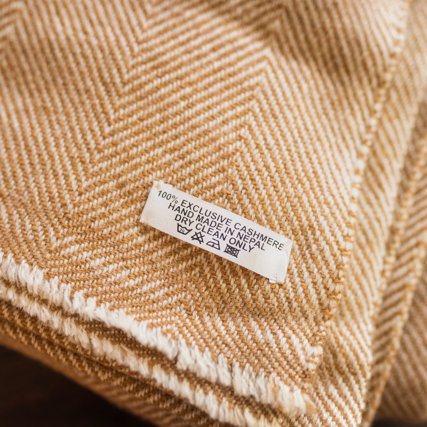 Auf dem Bild ist eine braune Kaschmirdecke zu sehen, die ein feines Fischgrätenmuster aufweist. Auf der Decke befindet sich ein weißes Etikett mit der Aufschrift „100% EXCLUSIVE CASHMERE, HAND MADE IN NEPAL, DRY CLEAN ONLY“ sowie vier Pflegesymbole. Der Deckenrand ist leicht fransig. Die Textur und das Muster des Stoffes sind deutlich sichtbar und wirken hochwertig und weich.